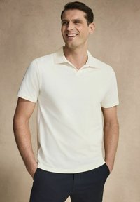 Kortsleeved polo tröja i off-white bomull med klassisk krage och mjuk textur. Bärs avslappnat med marinblå byxor.