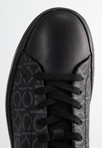 Baskets noires avec un motif texturé, un bout lisse noir et des lacets noirs. Le design est moderne avec un détail de logo subtil sur le côté.