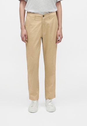 Person iført beige bukser med lige ben, hvide sneakers og en lysegrå kortærmet skjorte, stående mod en ensfarvet hvid baggrund.