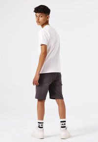 Camiseta blanca de manga corta, pantalones cortos grises, zapatillas blancas con detalles en negro y calcetines a rayas. El modelo está posicionado de espaldas.