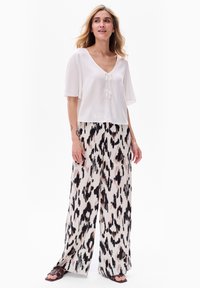 Biała bluzka z krótkim rękawem, z dekoltem w serek i detalem związanym, sparowana z szerokimi spodniami w czarno-beżowy abstrakcyjny wzór. Płaskie sandały.