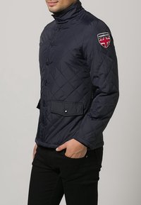 Gequiltete dunkelblaue Jacke mit Stehkragen, Knopfverschluss und Seitentasche. Mit einem Aufnäher im Design der Union Jack auf der Schulter.
