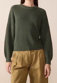 Pull vert en tricot avec manches longues et texture côtelée, associé à un pantalon plissé marron clair. Encolure ronde simple et poignets ajustés.