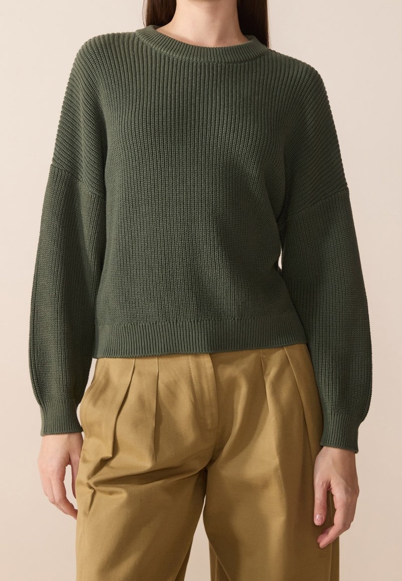 Pull vert en tricot avec manches longues et texture côtelée, associé à un pantalon plissé marron clair. Encolure ronde simple et poignets ajustés.