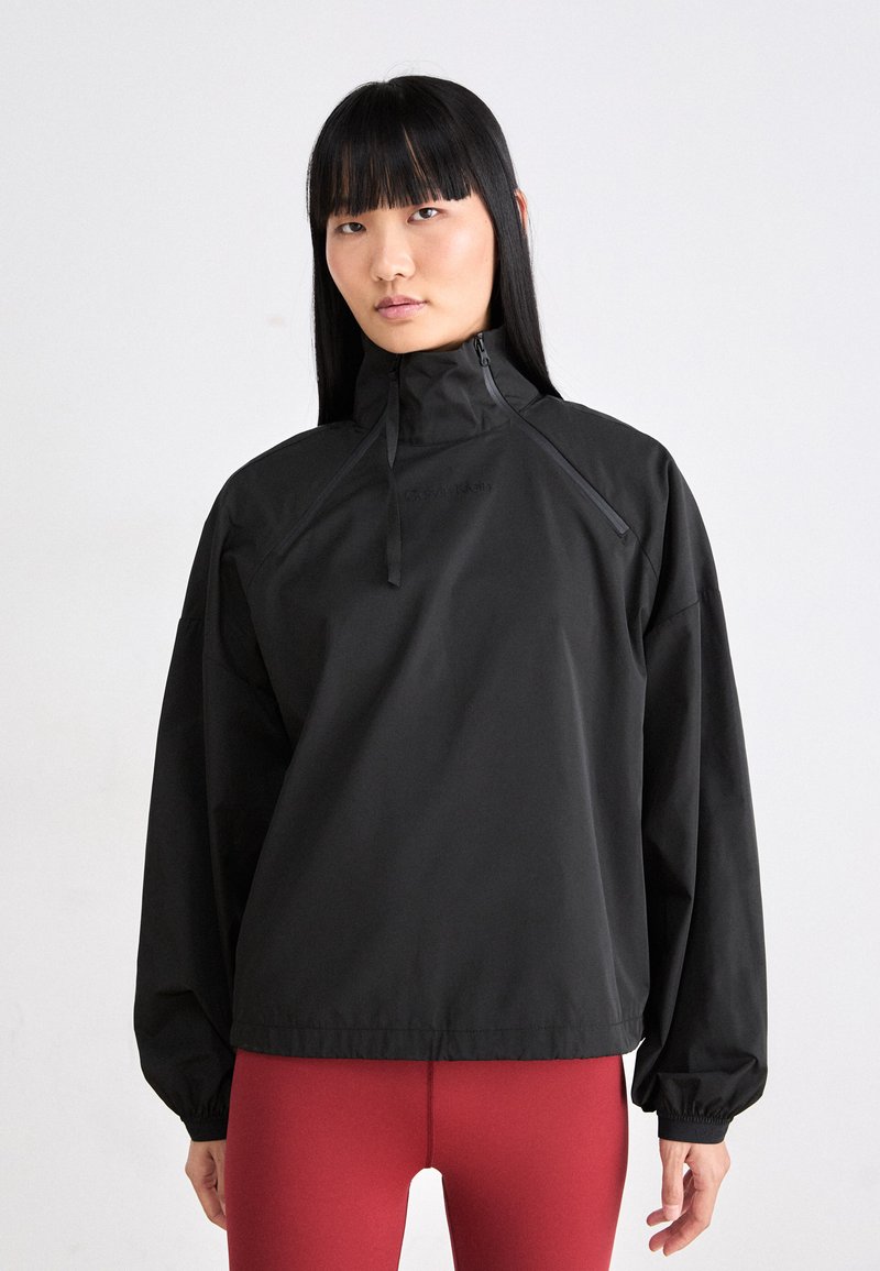 Schwarze leichte Pulloverjacke mit hohem Kragen und halbem Reißverschluss. Lange Ärmel mit elastischen Bündchen und lockerer Passform. Glatte, matte Textur.