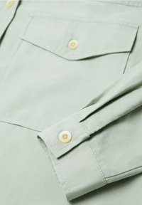 Chemise en coton vert clair avec une poche poitrine, une patte de boutonnage, et des boutons jaunes sur les poignets et le devant. Texture lisse.