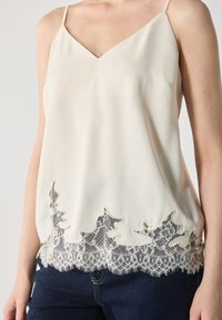 Camisole en satin crème avec des fines bretelles, ornée de délicates garnitures en dentelle noire au niveau de l'ourlet, créant un bord en forme de scallop et un contraste texturé.