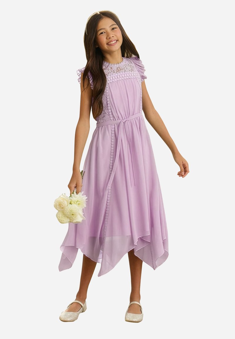 Niña sonriente con un vestido de encaje lila con cinturón, sosteniendo flores blancas y llevando zapatos brillantes y una diadema.