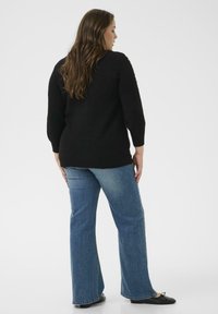 Schwarzer, strukturierter Strickpullover mit Dreiviertelärmeln, kombiniert mit weit geschnittenen blauen Jeans. Die Schuhe sind schwarz, flach und mit einer Schleifendetails.