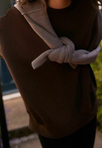 Brown sweatshirt with a crew neck, paired with a light gray knit scarf tied at the neck, creating texture contrast against the smooth fabric.