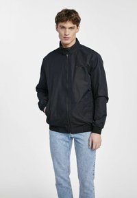 Czarna kurtka na zamek z wysokim kołnierzem, elastycznymi mankietami i dołem. Posiada subtelne czerwone akcenty na klatce piersiowej i kieszeniach. Noszona z jasnoniebieskimi dżinsami.