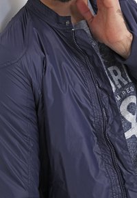 Marinblå nylon bomberjacka med hög krage, framzipper och ribbade detaljer. Texturerad yta med lätt glans.