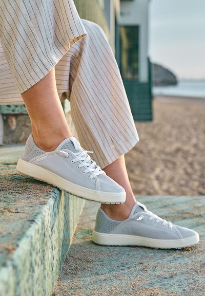 Person wearing light gray lace-up sneakers and striped pants sitting on patterned stone step near a sandy beach.