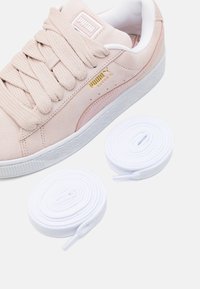 Puma mockasiner i ljusrosa med texturerad ovandel, vit gummisula och gyllene logotypdetaljer. Två vita skosnören bredvid skorna.