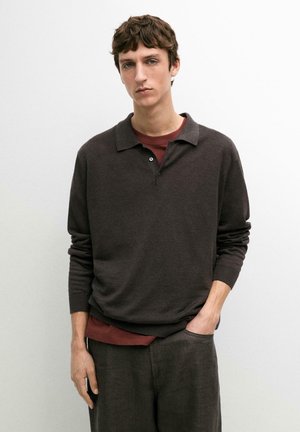 Ung mand med kort brunt hår iført en mørkebrun langærmet polo og en bordeaux skjorte, stående med den ene hånd i lommen.