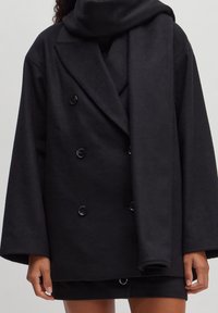 Musta kaksirivinen takki pehmeästä, teksturoidusta kankaasta, jossa suuret napit ja leveät hihat, yhdistettynä vastaavaan huiviin.