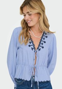 Blauwe en witte gestreepte blouse met lange mouwen, gegolfde zoom, geborduurde marineblauwe accenten en een striksluiting bij de taille.