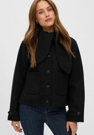 Femme aux longs cheveux châtain clair portant une veste noire boutonnée avec de grandes poches et une écharpe assortie, associée à un jean bleu.