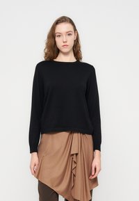 Μαύρο πλεκτό πουλόβερ με μακριά μανίκια και ριμπ λαιμόκοψη, φορεμένο πάνω από ανοιχτό καφέ, ασύμμετρη φούστα τύπου wrap. Ομαλή υφή και άνετη εφαρμογή.