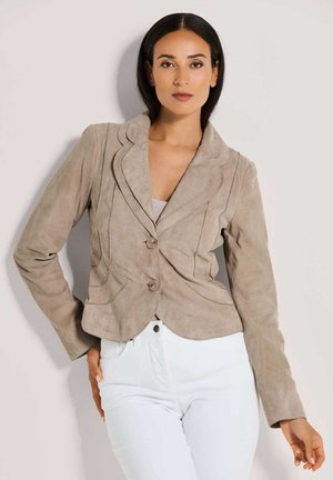 Helles taupe Suede-Blazer mit Reverskragen, zwei Knöpfen, abgerundetem Saum und genähten Details, kombiniert mit weißen hochgeschnittenen Hosen.