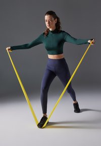 Fitness vježba u zelenoj majici dugih rukava i tamnoplavim legginzima, koristeći žuti otporni trak. Pozadina je siva s mekim osvjetljenjem.