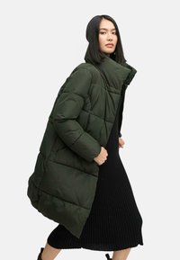 Long, puffy coat in dark green with quilted sections. High collar and open front. Worn over a black ribbed sweater dress. Smooth texture.