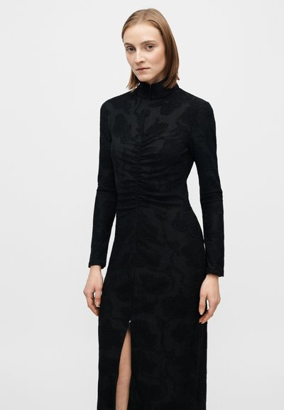 Fekete hosszú ujjú ruha, amely texturált virágmintákkal, magas nyakkal és elöl hasítékkal rendelkezik. Az anyag selymesnek tűnik, szabása testhezálló.