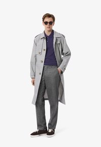 Ljusgrå rutigt trench coat med guldfärgad knappar över en mörklila polo, kombinerat med grå byxor och bruna sneakers.