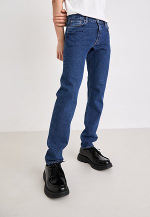 Osoba nosząca niebieskie jeansy z denimu i czarne skórzane buty sznurowane, stojąca na jasnej podłodze na jasnym tle.