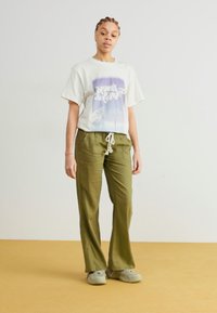 White t-shirt with graphic print, loose green pants with drawstring waist, and grey shoes, standing on a yellow floor against a light background.