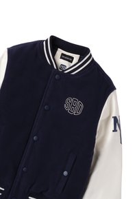Granatowa kurtka varsity z białymi skórzanymi rękawami, prążkowanym kołnierzem i zatrzaskami. Posiada haft "SBD" i "NY" na rękawie.