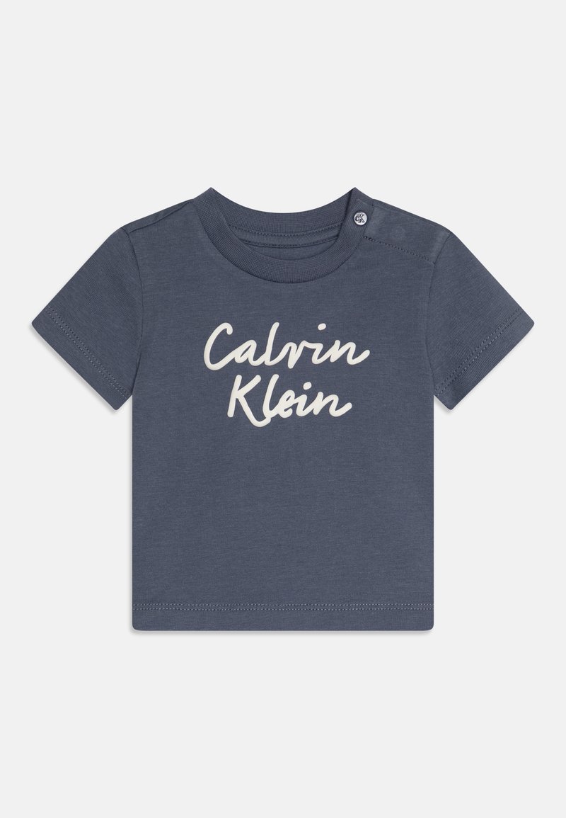 Marineblå bomulls t-skjorte med korte ermer som har "Calvin Klein" i hvit kursiv tekst, med en trykknapp på skulderen for enkel påkledning.