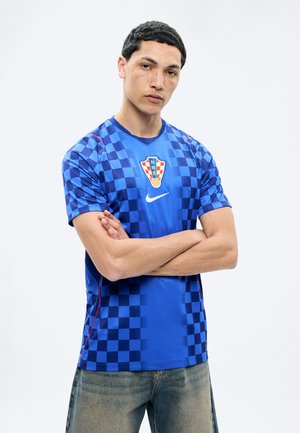 Noor mees lühikeste juuste ja pigmenteeritud näoga seisab käed rüpes, kandes sinise ruudulise Horvaatia jalgpallimängija särgi ja kulunud teksaseid.