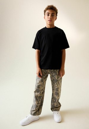 Chlapec s kučeravými vlasmi stojí, má na sebe čierne tričko, široké nohavice s leopardím vzorom a biele tenisky na jednofarebnom pozadí.