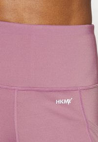 Jasnofioletowe legginsy z wysokim stanem, z widocznymi detalami przeszycia. Ozdobione haftowanym białym logo na pasie. Miękka, gładka tekstura.