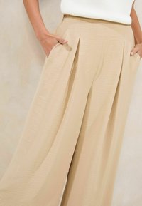 Pantalons plissés beige en tissu texturé, avec des poches latérales et une coupe ample. La ceinture est lisse, ce qui améliore le confort et le style.