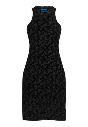 Femme portant une robe noire en dentelle sans manches avec une fente et une veste en cuir noire drapée sur ses épaules, debout pieds nus.