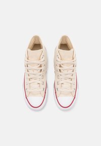 Hoge sneakers van crèmekleurig canvas met een witte rubberen neus en zool. Heeft rode accenten langs de zool en beige veters door zilveren oogjes.