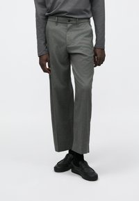 Szürke nadrág texturált anyagból, egyenes szárú kialakítással és jól látható elülső zsebekkel, fekete cipővel és bokaig érő zoknival párosítva.