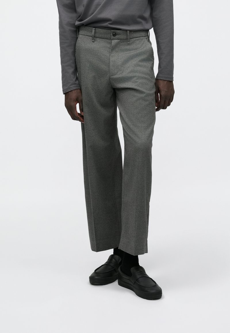 Szürke nadrág texturált anyagból, egyenes szárú kialakítással és jól látható elülső zsebekkel, fekete cipővel és bokaig érő zoknival párosítva.