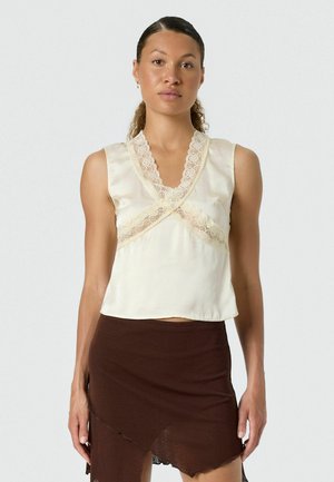 Vrouw draagt een mouwloze crèmekleurige blouse met kanten rand en asymmetrische donkerbruine rok, staand tegen een eenvoudige lichte achtergrond.