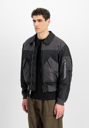Junger Mann mit kurzen lockigen Haaren, der eine schwarze und dunkelgraue Bomberjacke mit großen Fronttaschen und olivgrünen Hosen trägt.