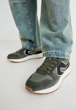 Zapatillas deportivas verdes con materiales de ante y textil, que presentan detalles en blanco y negro, cordones y una suela de goma. Combinadas con unos jeans holgados de color azul claro.