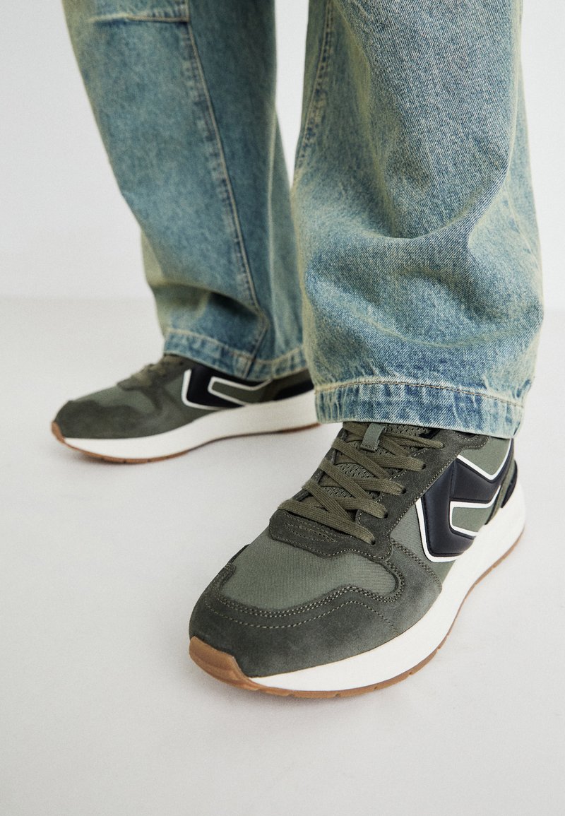 Zapatillas deportivas verdes con materiales de ante y textil, que presentan detalles en blanco y negro, cordones y una suela de goma. Combinadas con unos jeans holgados de color azul claro.