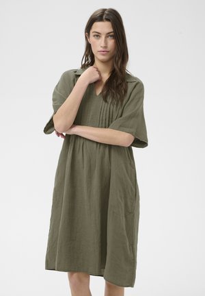 Young woman with long brown hair wearing loose olive green knee-length dress with short sleeves, standing against plain white background.