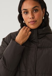 Bruine pufferjack met een hoge kraag, voorzien van gestikte secties en een trekkoord bij de nek. Glad, glanzend oppervlak. Metallic hardware accenten.