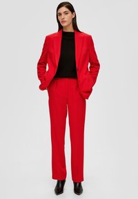 Czerwony żakiet dopasowany i pasujące do niego spodnie z gładkiego materiału, połączone z czarnym swetrem o prążkowanej fakturze, uzupełnione czarnymi, szpiczastymi botkami za kostkę.