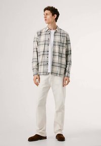 Camisa xadrez de flanela em cinza e branco, design com botões na frente, usada sobre uma t-shirt branca, combinada com calças bege e sapatos castanhos.