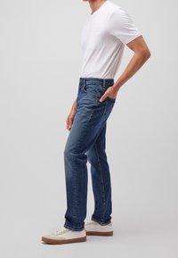 Hombre con una camiseta blanca de manga corta, jeans de mezclilla azules con la mano en el bolsillo y zapatillas blancas con suelas de goma, de pie de lado.