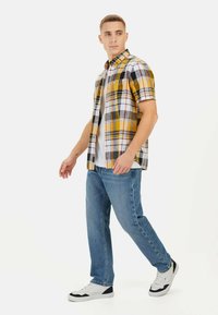 Korte mouw geruit overhemd in geel, zwart en wit, gecombineerd met blauwe jeans en witte sneakers. Het overhemd heeft een button-down kraag.